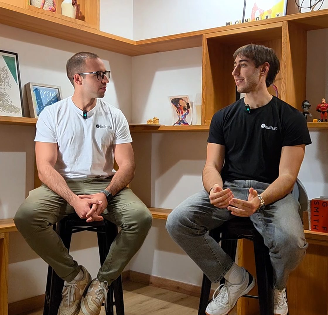 Los dos socios de DoGeo sentados en taburetes, de perfil y hablando entre ellos, con micrófonos de solapa y camisetas de Kulturo, en una sala con estanterías de madera.
